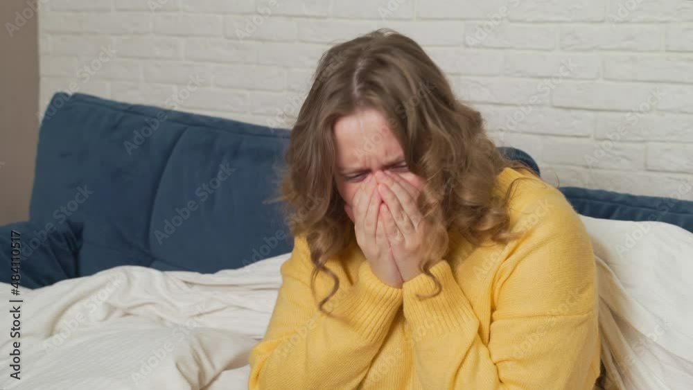 Woman in sweater sits on bed and coughs and sneezes. She has cold, flu, coronavirus. Her throat hurts. She has fever and she wraps herself in blanket. Flu, cold, coronavirus epidemic isolation