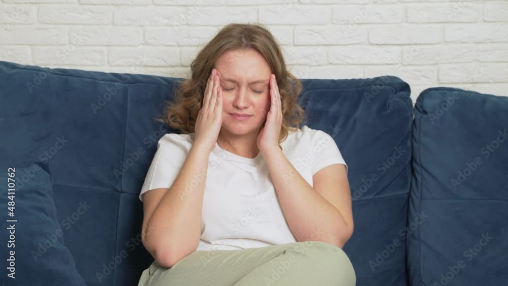 Young woman is sitting on the bed, she has migraine. She is massaging the temples of her head due to severe spasms. Suffering on face, she is breathing deeply. Cold, flu, migraine, epidemic, cramps