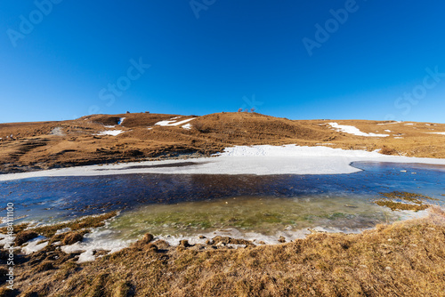 Winter landscape of Lessinia Plateau Regional Natural Park (Altopiano della Lessinia) with a small frozen pond for cows and brown pastures. Erbezzo municipality, Verona province, Veneto, Italy, Europe