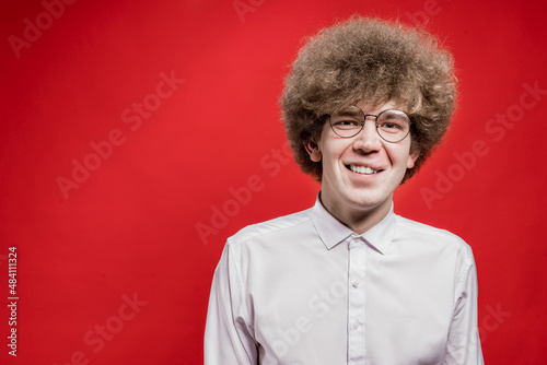 A handsome curly-haired man in a shirt and glasses smiles at the camera on a red background. Unusual hairstyle. The man has poor eyesight © watman