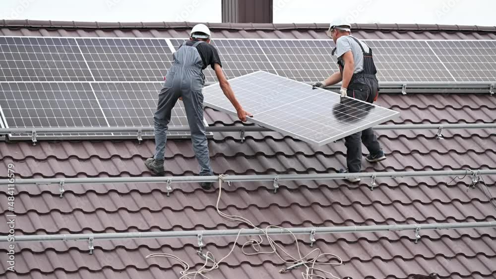 Men workers lifting up photovoltaic solar modul on roof of house. Electricians in helmets installing solar panel system outdoors. Concept of alternative and renewable energy.