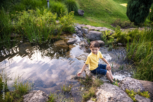 A young boy sits on the river bank, view from top. Warm summer or spring day. Cute Caucasian kid play with water