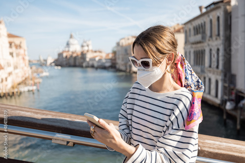 Young woman in medical mask traveling during pandemic in Italy. Concept of social rules of wearing a mask during a pandemic. Woman using smartphone while standing on the bridge in Venice