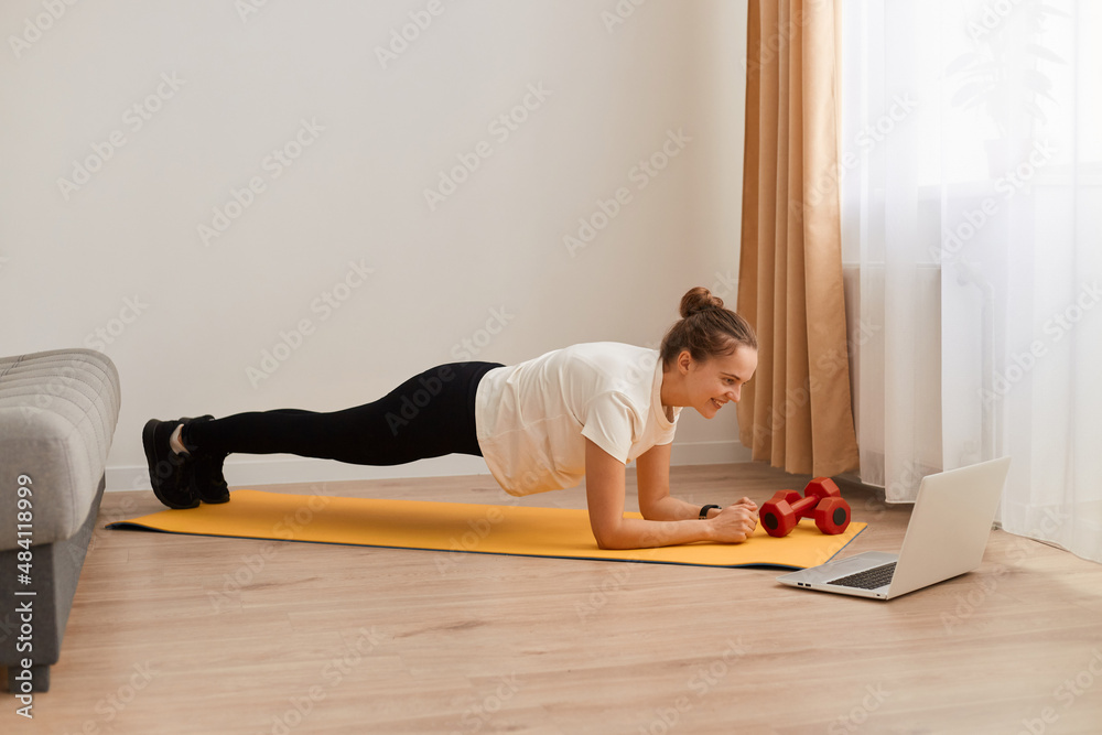 Full length side view portrait of athletic woman wearing white T-shirt and black leggins training her abdominal muscles, standing in plank pose, fitness at home on yoga mat. Full length side view portrait of athletic woman wearing white T-shirt and black leggins training her abdominal muscles, standing in plank pose, fitness at home on yoga mat.