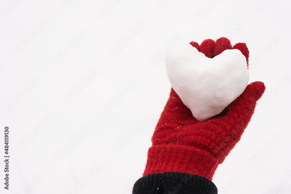 Close-up view stock photography of small white snow heart in hand of woman. Female hand in warm red glove holding cute snowy heart. Love to winter season, St. Valentine's Day, Christmas concept
