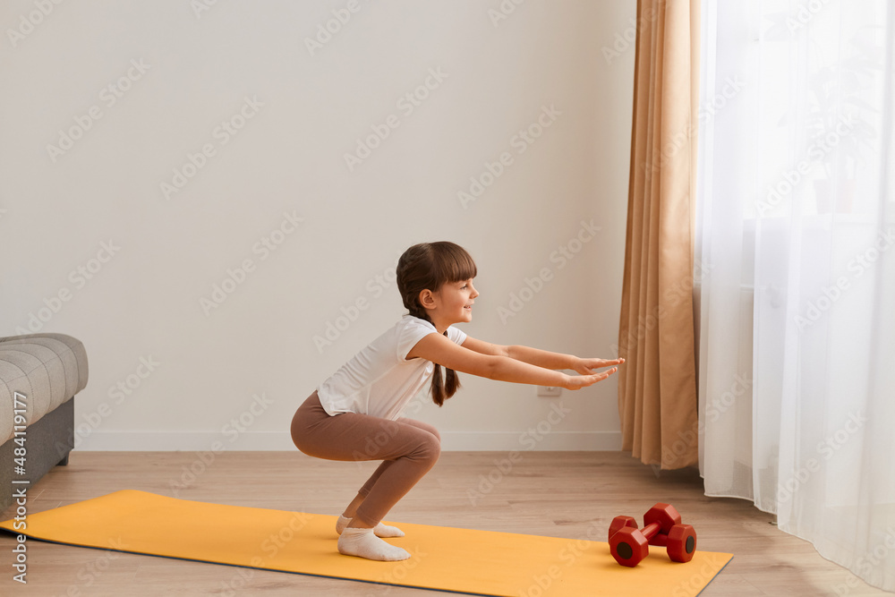 Portrait of little girl doing squat exercises workout at home, cute kid training on a mat indoor, dark-haired female kid having exercises near the window in her room. Portrait of little girl doing squat exercises workout at home, cute kid training on a mat indoor, dark-haired female kid having exercises near the window in her room.
