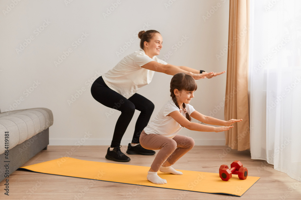Mother and little child daughter practicing are engaged in fitness, sport exercise mat at home, woman with child doing squatting together with outstretched hands.