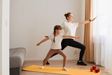 Indoor shot of beautiful young woman and her charming little daughter are smiling while doing yoga together at home, standing on yoga mat, doing warrior pose.
