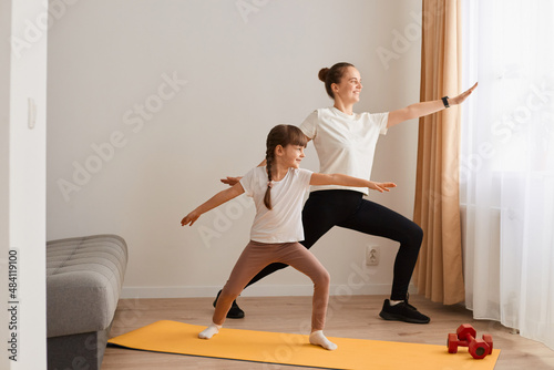 Indoor shot of beautiful young woman and her charming little daughter are smiling while doing yoga together at home, standing on yoga mat, doing warrior pose.