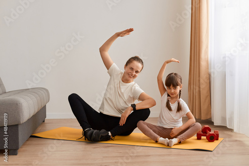 Indoor shot of slim woman with hair bun sitting in lotus position on mat practicing yoga with her cute female child, bending to side with raised hand, doing stretching workouts with daughter.