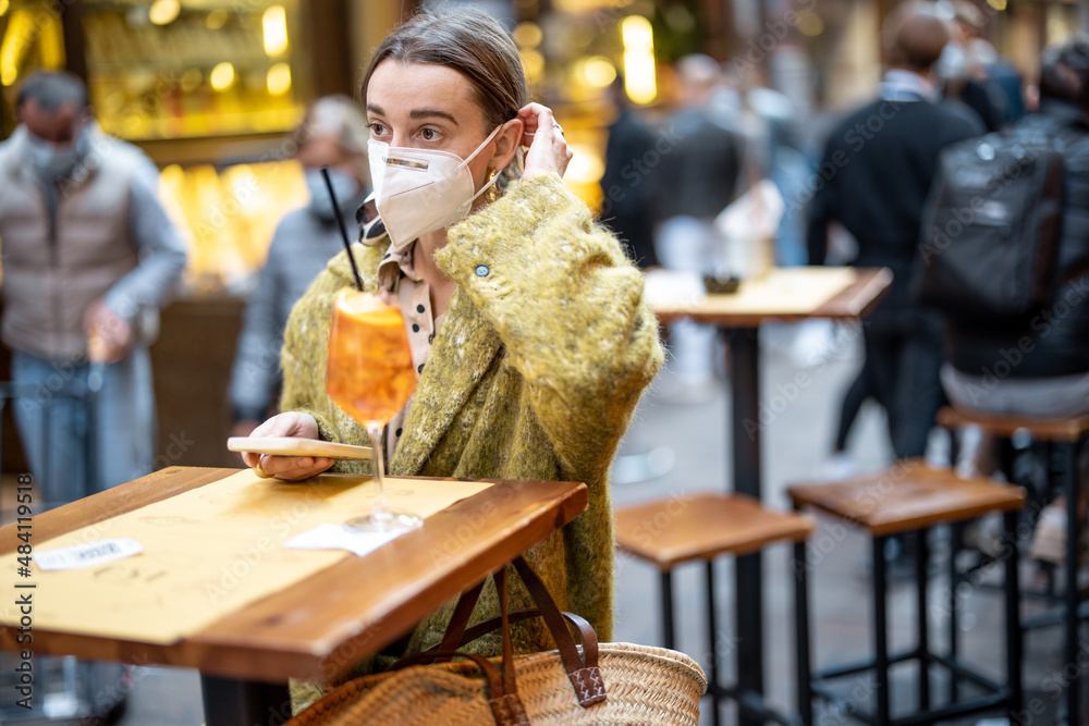 Woman takes off medical mask while sitting with alcoholic cocktail at outdoor cafe on the street in Bologna. Concept of Italian gastronomic culture and new social rules related to pandemic