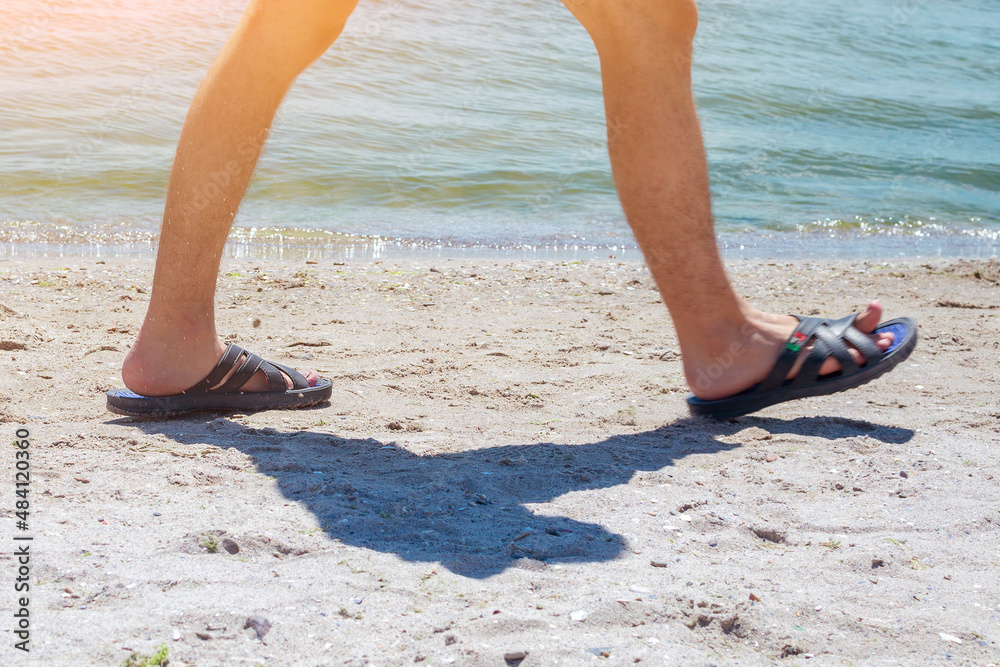 Thin and hairy male legs in slippers. A young man walks in sunny weather along the beach along the sea coastline. In the frame only legs