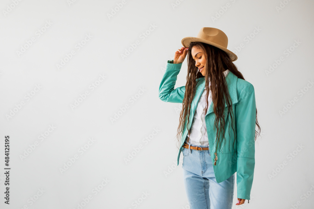 Sincere cheerful dark-skinned teenager or hipster girl is dressed in a turquoise leather jacket and a beige hat, posing cheerfully in a spacious bright studio Sincere cheerful dark-skinned teenager or hipster girl is dressed in a turquoise leather jacket and a beige hat, posing cheerfully in a spacious bright studio