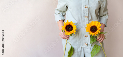 A girl in a light green cotton jumpsuit holds two sunflowers in her hands, a beige monochrome background. The concept of an ecological lifestyle and clothing. copy space