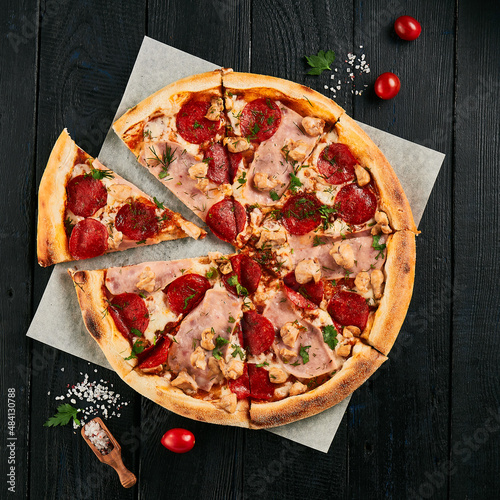 American meat pizza on dark wood background. Italian dish - homemade pizza with ham, salami, pepperoni and chicken on parchment. Junk food on wooden table. Pepperoni pizza in rustic style.