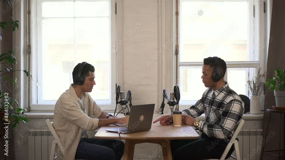 Two male speakers talking and recording podcast at table with laptop in modern studio spbd. Young men talk and look with smiles, record audio and sit at desk in front of microphones in interior. Guest Two male speakers talking and recording podcast at table with laptop in modern studio spbd. Young men talk and look with smiles, record audio and sit at desk in front of microphones in interior. Guest