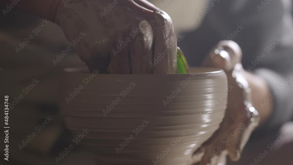 A potter sculpts soft clay with wet hands, creating handmade ceramic creative pot, rotating on potter's wheel. Clay shaping and sculpting, close up hand creative work in working workshop. Slow motion.