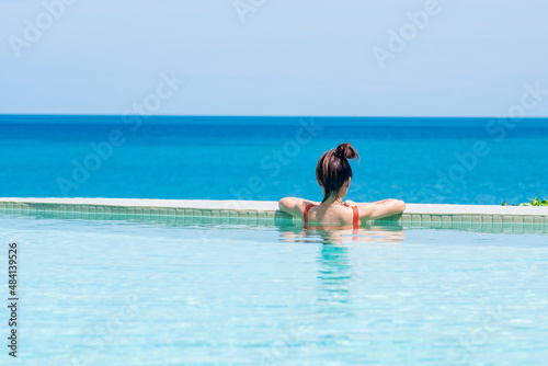 Happy woman in orange swimsuit swimming in infinity pool at luxury hotel against ocean front. young female enjoy in tropical resort. Relaxing, summer, travel, holiday, vacation and weekend concept