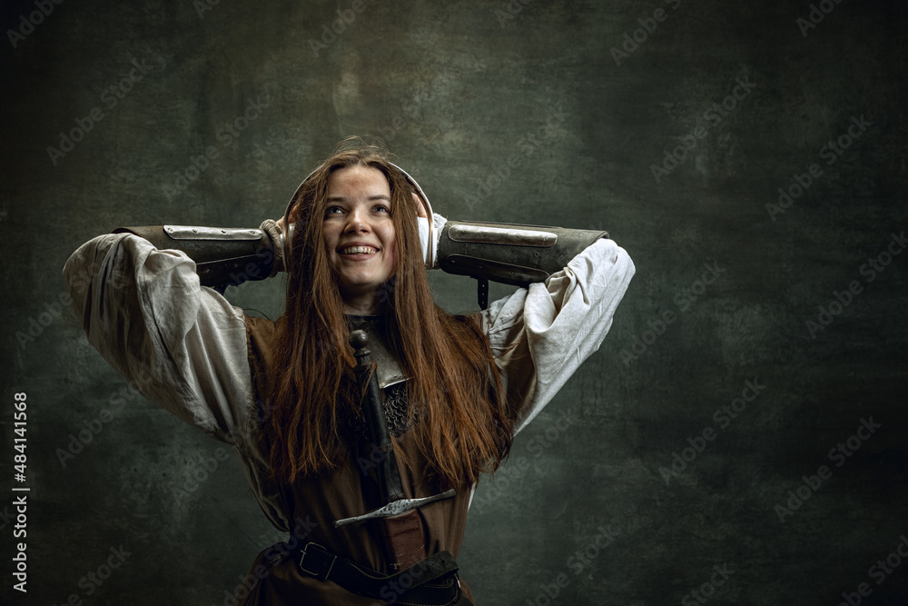 Young medieval warrior or knight with dirty wounded face in headphones listening to music isolated over dark vintage background. Comparison of eras, history Young medieval warrior or knight with dirty wounded face in headphones listening to music isolated over dark vintage background. Comparison of eras, history