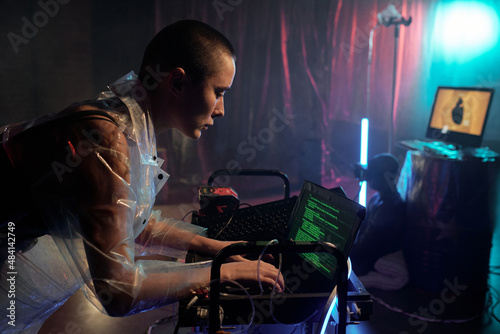 Young serious female in cellophane coat decoding data and looking at laptop display against male cyberpunk sitting on the floor