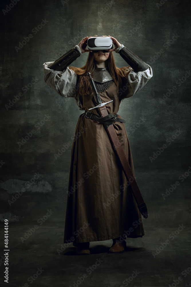 Portrait of young wman, medieval warrior, knight with long hair in VR headset isolated over dark vintage background. Comparison of eras, history Portrait of young wman, medieval warrior, knight with long hair in VR headset isolated over dark vintage background. Comparison of eras, history