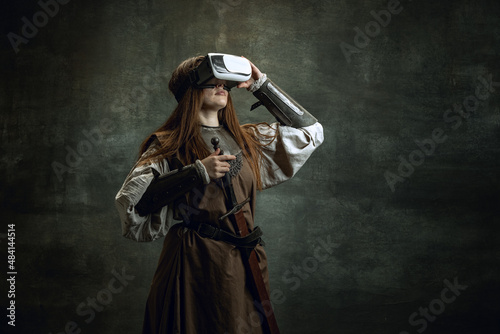 Portrait of young wman, medieval warrior, knight with long hair in VR headset isolated over dark vintage background. Comparison of eras, history photo