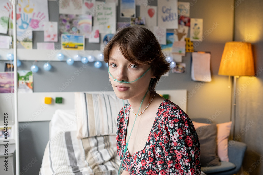 Portrait of sick teenage girl breathing with oxygen tube looking at camera while sitting on hospital bed at the ward Portrait of sick teenage girl breathing with oxygen tube looking at camera while sitting on hospital bed at the ward