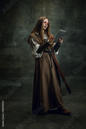 Vintage portrait of young beautiful girl in image of medieval warlike woman using modern gadget isolated over dark background. Comparison of eras, history