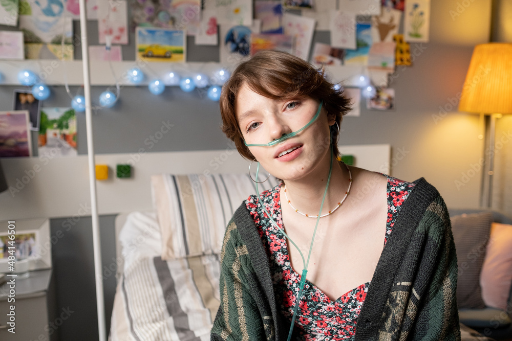 Portrait of teenage girl breathing with special oxygen tube looking at camera while sitting on bed at the ward Portrait of teenage girl breathing with special oxygen tube looking at camera while sitting on bed at the ward