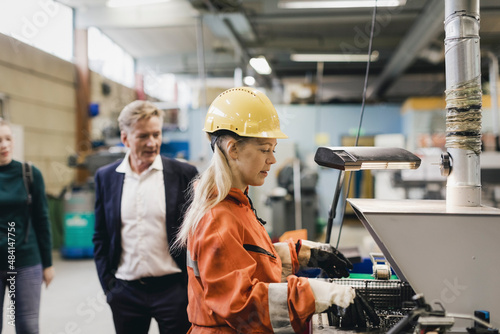 Female industrial worker in hardhat working on machinery while managers in background at factory photo