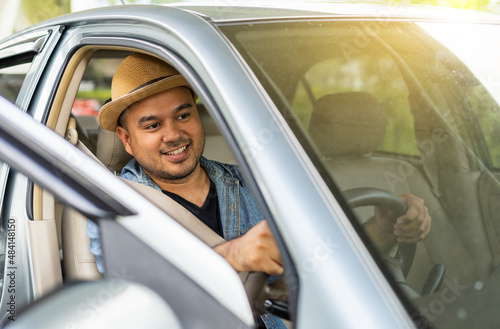 Happy male driver smiling while sitting in a car with open front window. Young asian man smile and looking through car window. View of a Young man in hat driving his car to travel.