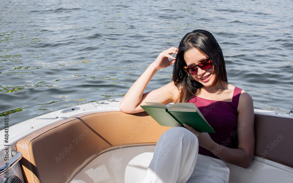Asian beautiful adorable young woman reading book, wearing casual shirt and sunglasses, sitting on boat or ship for traveling outdoor in summer time, smiling with happiness. Lifestyle concept.