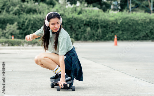Beautiful sportive hipster happy Asian woman wearing casual shirt and shorts with headphone to listen music while playing skateboard as hobby in free time day on holiday, smiling at outdoor park.