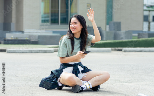 Beautiful happy Asian extreme hipster woman smiling, talking mobile phone and waving hand for greeting, sitting on skateboard, relax playing outdoor activity on holidays in summer vacation.