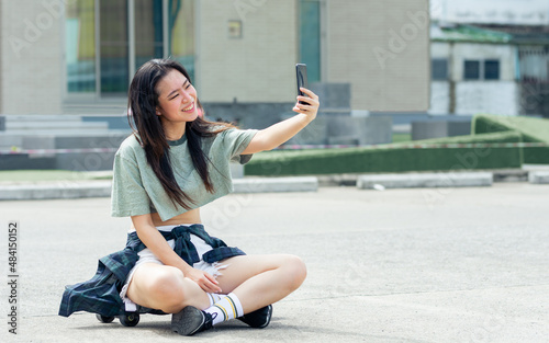 Beautiful happy Asian extreme hipster woman smiling, using mobile phone and taking selfie photo alone, sitting on skateboard, relax playing outdoor activity on holidays in summer vacation.
