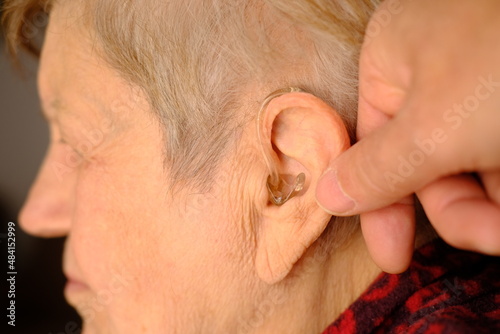 Close-up view of hands inserts hearing aid to the elderly woman's ear. Senior woman with symptoms of hearing loss wears a hearing aid device to hear properly. 