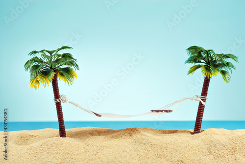 Fototapeta Naklejka Na Ścianę i Meble -  Miniature hammock on artificial palm trees. Layout of the beach with palm trees in the sand. Blue sky in the background. The concept of rest and travel.