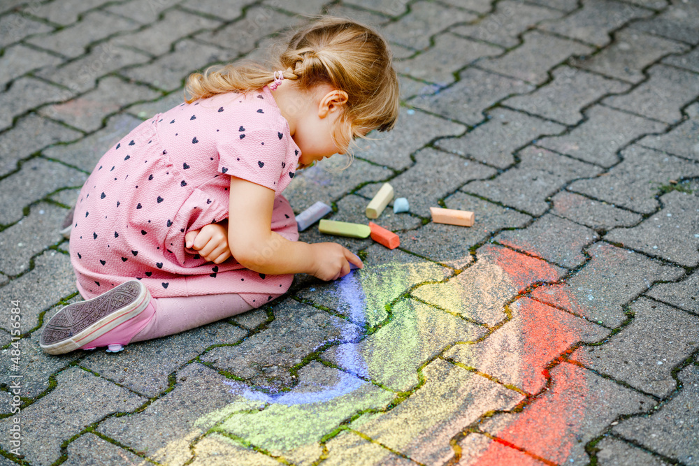 Little preschool girl painting rainbow with colorful chalks on ground on backyard. Positive happy toddler child drawing and creating pictures. Creative outdoors activity in summer. Little preschool girl painting rainbow with colorful chalks on ground on backyard. Positive happy toddler child drawing and creating pictures. Creative outdoors activity in summer.