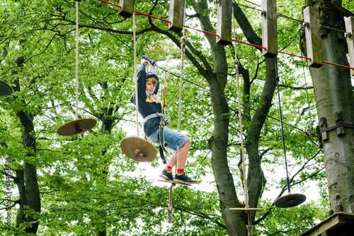 School boy in forest adventure park. Acitve child, kid in helmet climbs on high rope trail. Agility skills and climbing outdoor amusement center for children. Outdoors activity for kid and families.