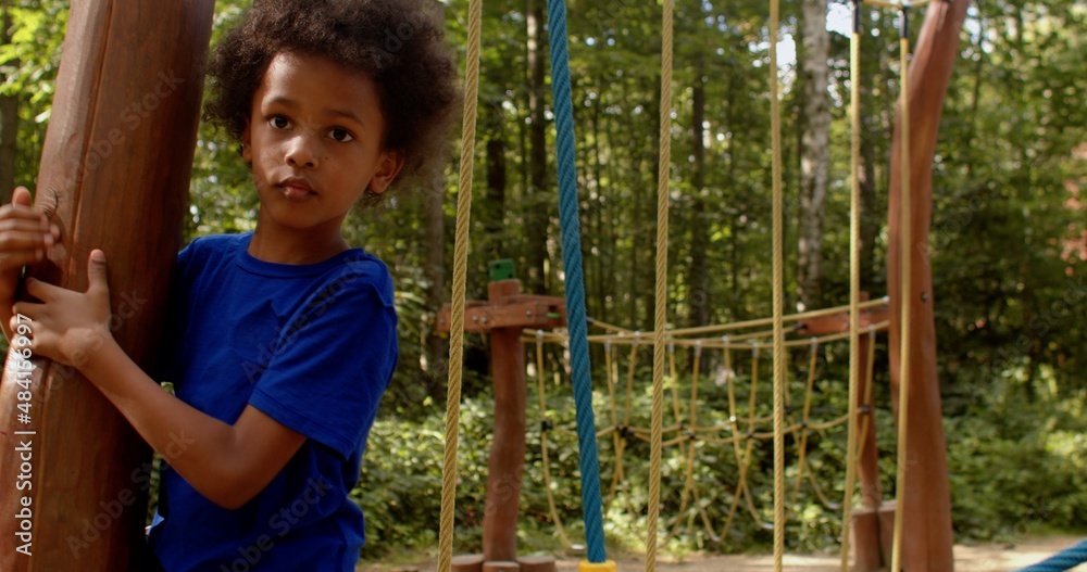 Obraz premium The boy deftly moves along the hinged rope path on the playground, tenaciously grabbing the ropes with his hands. Harmonious development of children's body through outdoor activities.