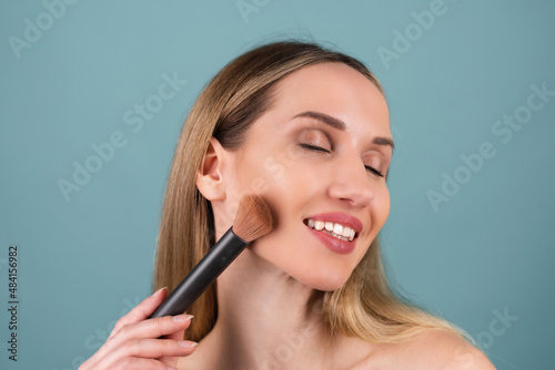 Close-up beauty portrait of topless woman with perfect skin and natural makeup, full nude lips, holding an blush brush for contouring