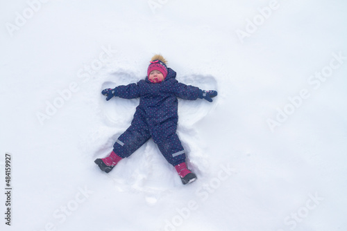 A Caucasian baby girl in a blue winter jumpsuit and a knitted hat lies on the snow and portrays an angel in winter
