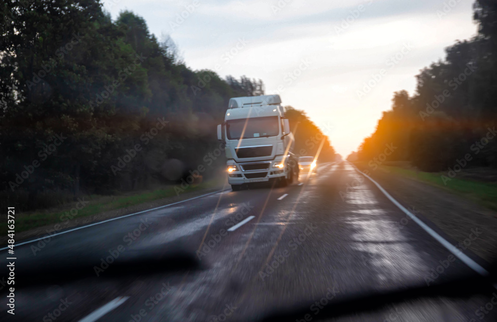 View from an oncoming car on a wet slippery road in the rain. Trucks with headlights on a sunset background. Aquaplaning on the road, poor visibility. Copy space for text, blurry