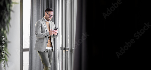 Smiling man in casuals standing in office. Businessman with mobile phone in hand. Smiling businessman using phone in office. Small business entrepreneur looking at his mobile phone and smiling.