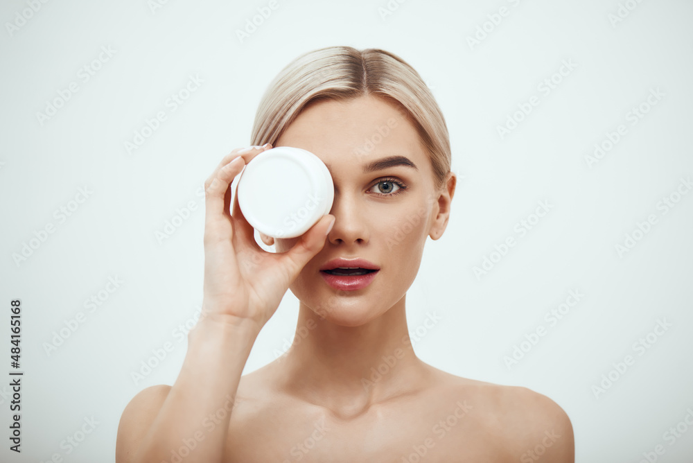 Deep hydration. Portrait of cute young blonde woman showing face cream while standing against grey background Deep hydration. Portrait of cute young blonde woman showing face cream while standing against grey background