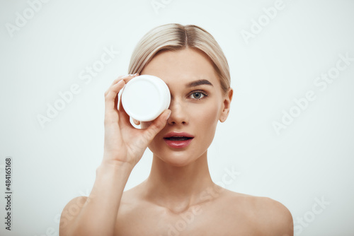Deep hydration. Portrait of cute young blonde woman showing face cream while standing against grey background