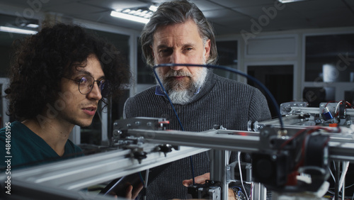 Male designer with curly hair showing and explaining work of 3D printer to mature bearded colleague during work in contemporary studio