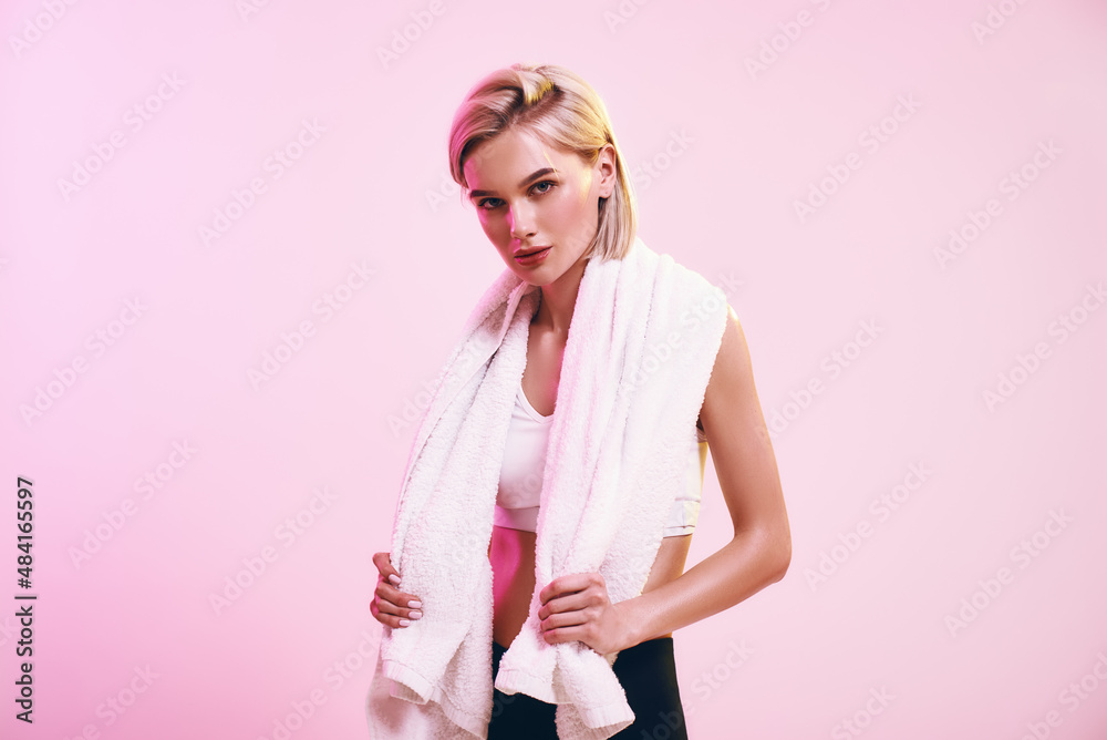 Time for rest. Attractive young blonde woman with towel on shoulders looking at camera while standing against pink background in studio
