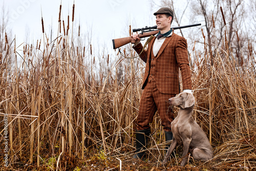 Hunting period, autumn season open. confident young caucasian hunter with dog and gun in brown trendy suit standing in autumn season nature, in search of trophy, prey. active sportive lifestyle. photo