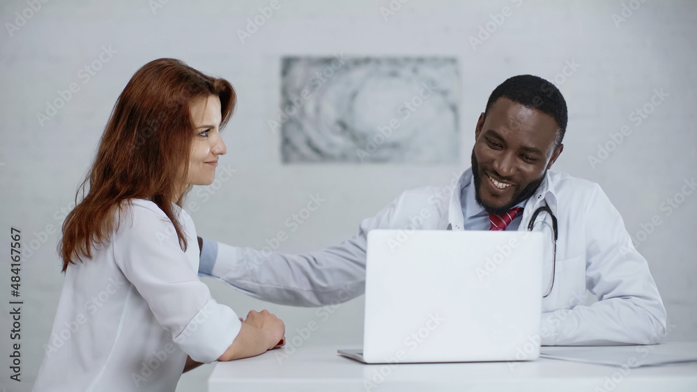 cheerful african american doctor talking with redhead patient near laptop on desk. cheerful african american doctor talking with redhead patient near laptop on desk.